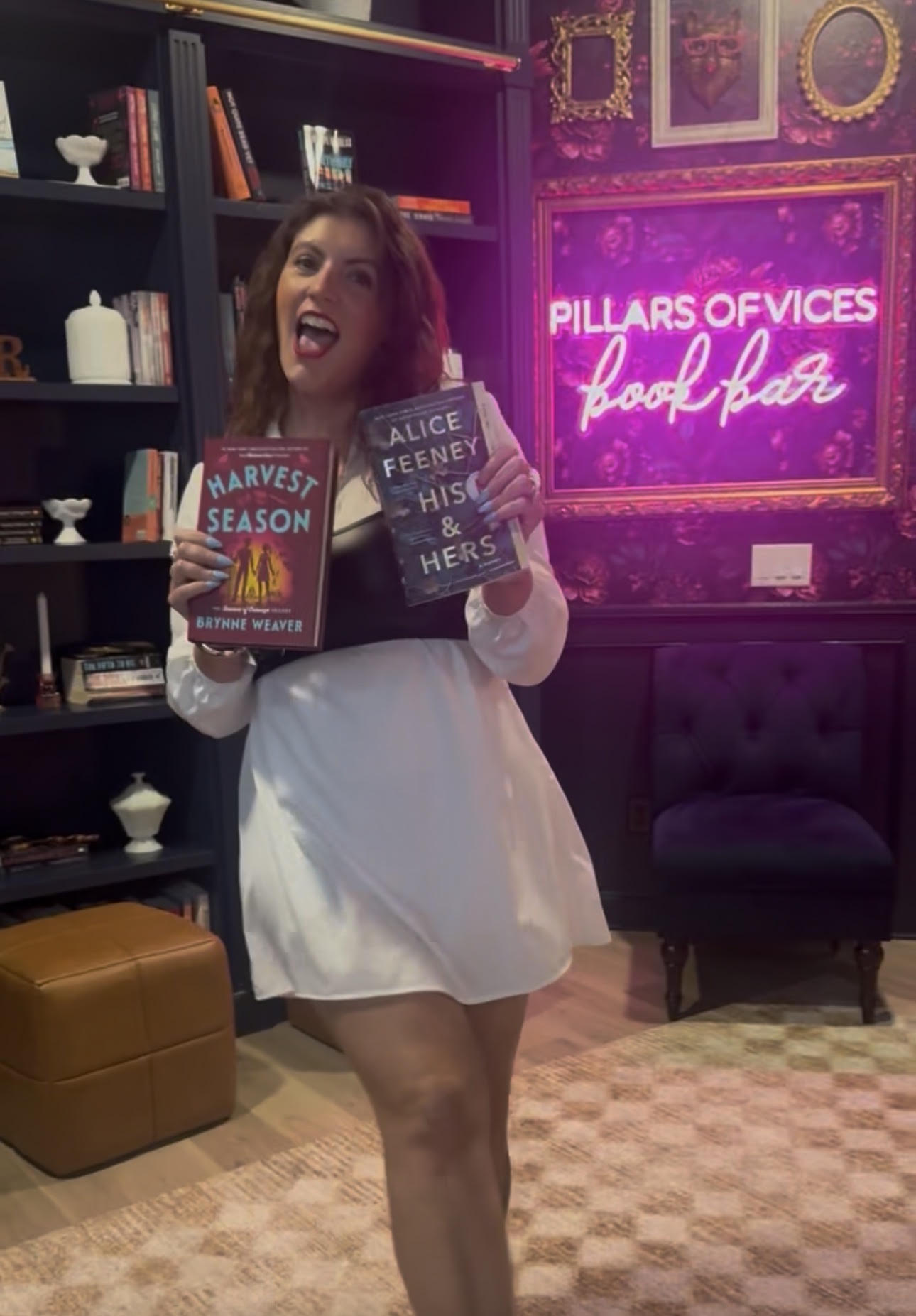 Jessica holding up August’s picks, Harvest Season and His & Hers, in front of the Pillars of Vices Book Bar sign