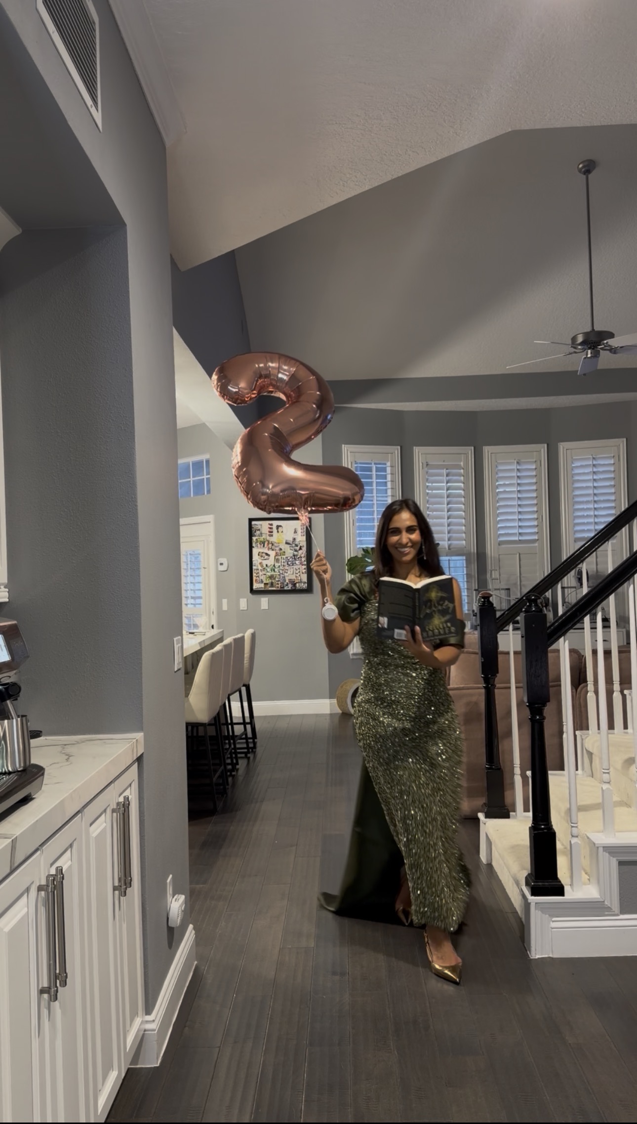 A Book Fetish Momma in a sparkly green gown holding a rose gold number 2 balloon and a copy of Verity