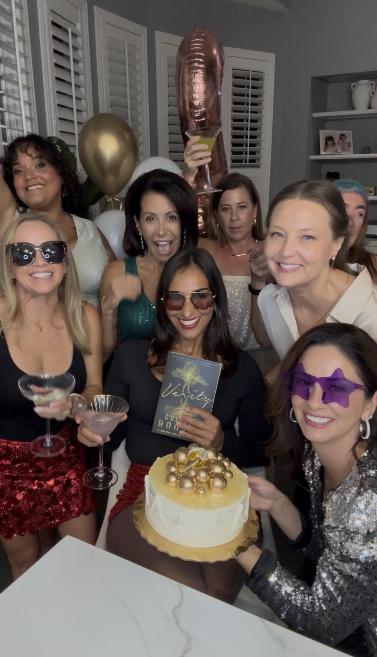 The Book Fetish Momma in their sparkly best, gathered around a gold anniversary cake with balloons and a copy of Verity